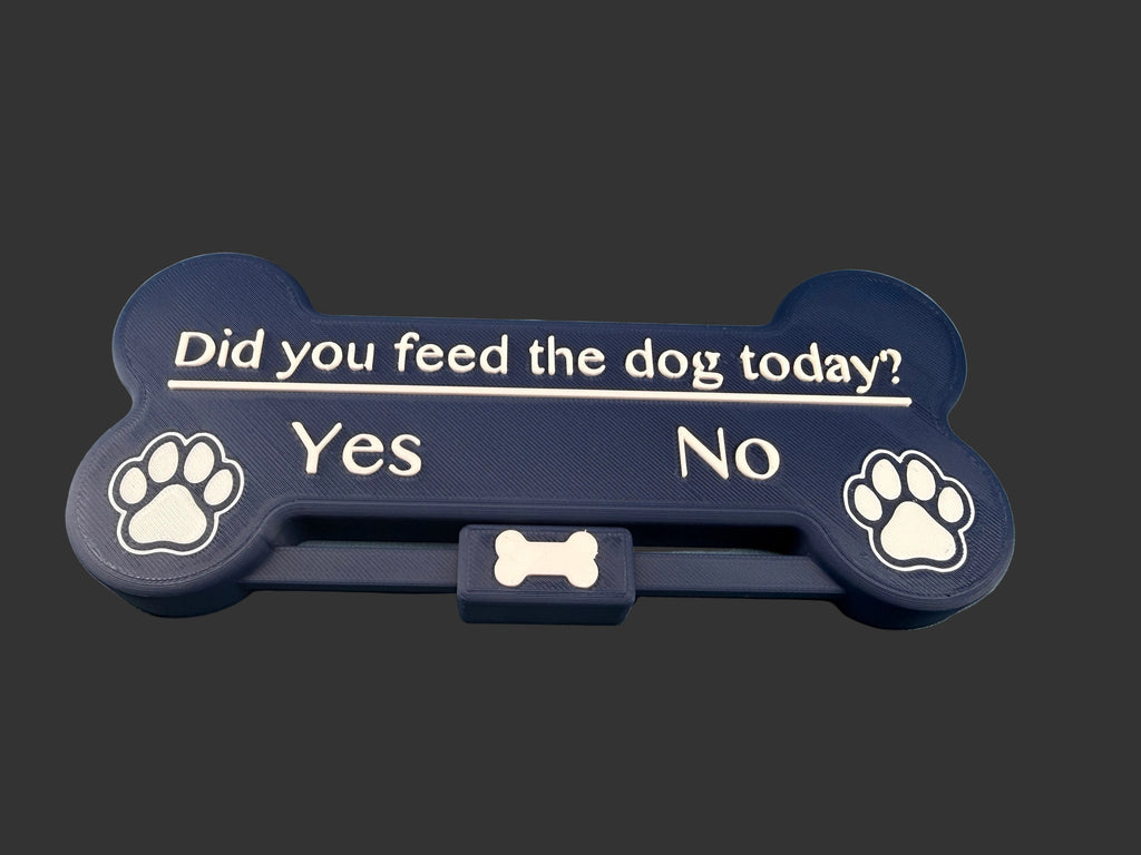 A bone-shaped pet feeder tray in dark navy blue with white text reading 'Did you feed the dog today? Yes No' and paw print icons on both ends, resting on a blue surface.