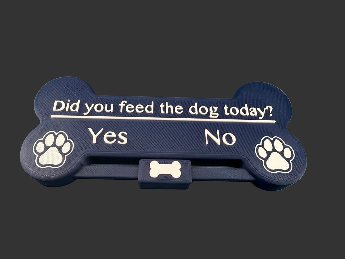 A bone-shaped pet feeder tray in dark navy blue with white text reading 'Did you feed the dog today? Yes No' and paw print icons on both ends, resting on a blue surface.